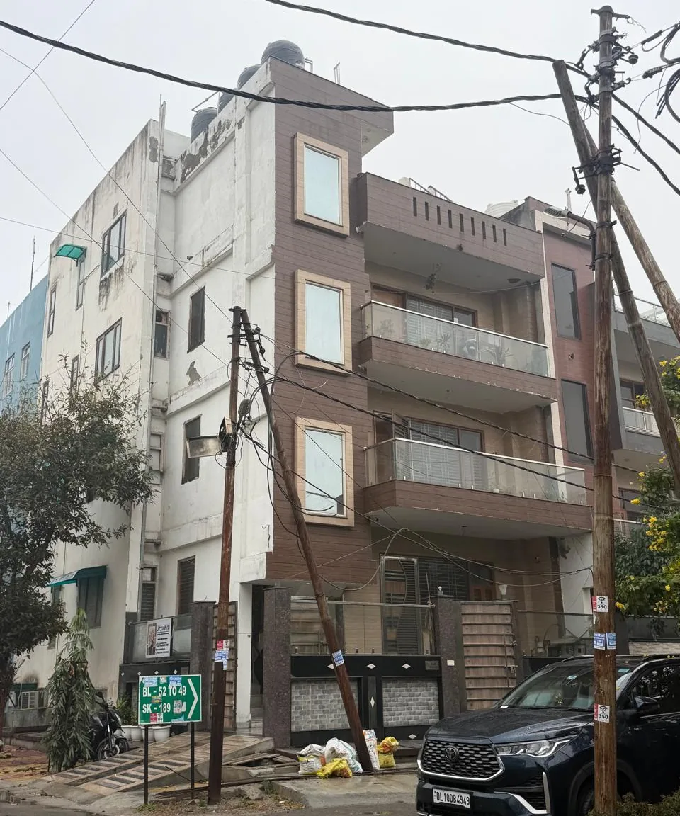 The same corner of the building as found on site: a white side wall and a brown-tiled front with glass balconies, overhead cables and a parked car.